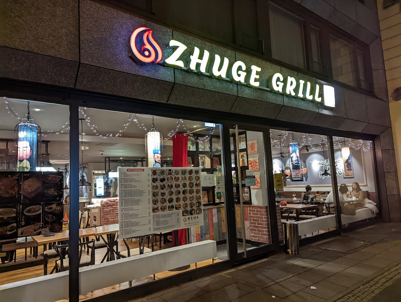 Außenansicht bei Nacht: leuchtender ZHUGE GRILL Schriftzug mit Flammen-Logo